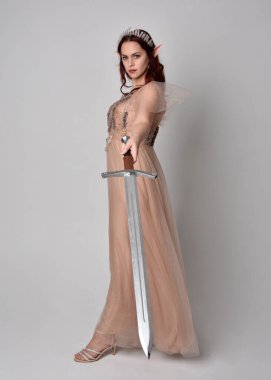  Kızıl saçlı, çekici fantezi tül cübbe ve taç giyen güzel bir kadın modelin tam boy portresi. Stüdyonun arka planında bir kılıç taşıyor..