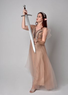  Kızıl saçlı, çekici fantezi tül cübbe ve taç giyen güzel bir kadın modelin tam boy portresi. Stüdyonun arka planında bir kılıç taşıyor..