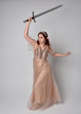  Kızıl saçlı, çekici fantezi tül cübbe ve taç giyen güzel bir kadın modelin tam boy portresi. Stüdyonun arka planında bir kılıç taşıyor..