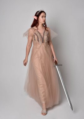  Kızıl saçlı, çekici fantezi tül cübbe ve taç giyen güzel bir kadın modelin tam boy portresi. Stüdyonun arka planında bir kılıç taşıyor..