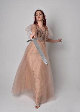  Kızıl saçlı, çekici fantezi tül cübbe ve taç giyen güzel bir kadın modelin tam boy portresi. Stüdyonun arka planında bir kılıç taşıyor..