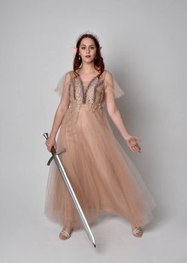  Kızıl saçlı, çekici fantezi tül cübbe ve taç giyen güzel bir kadın modelin tam boy portresi. Stüdyonun arka planında bir kılıç taşıyor..