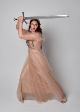  Kızıl saçlı, çekici fantezi tül cübbe ve taç giyen güzel bir kadın modelin tam boy portresi. Stüdyonun arka planında bir kılıç taşıyor..