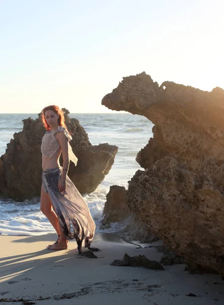 portrait of pretty female ship wrecked  model wearing  torn dress.  posing on the rocky  Ocean shoreline at sunset,
