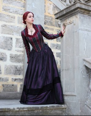 Tarihi Victoria tarzı bir elbise giyen kızıl saçlı bir kadının portresi taş mimariyle güzel bir yerde yürüyor..