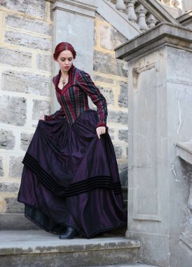 Tarihi Victoria tarzı bir elbise giyen kızıl saçlı bir kadının portresi taş mimariyle güzel bir yerde yürüyor..