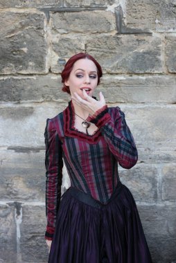 Tarihi Victoria tarzı bir elbise giyen kızıl saçlı bir kadının portresi taş mimariyle güzel bir yerde yürüyor..