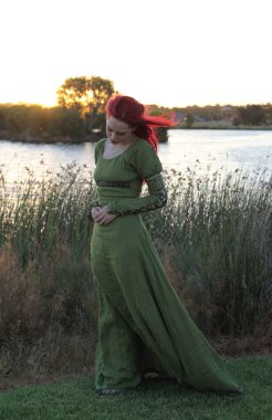 Ortaçağ fantezi elbisesi giymiş kızıl saçlı bir kadının tam boy portresi. Büyülü bir orman arka planında el hareketleriyle poz vermek.