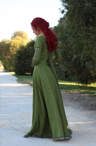 Полный портрет рыжеволосой женщины в красивом зеленом средневековом фэнтези-платье. Позирование с жестом руками на заколдованном фоне леса.