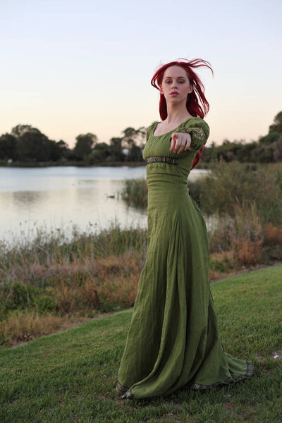 Полный портрет рыжеволосой женщины в красивом зеленом средневековом фэнтези-платье. Позирование с жестом руками на заколдованном фоне леса.