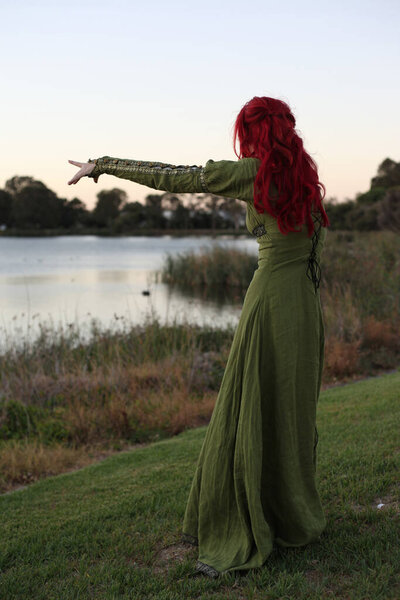 Полный портрет рыжеволосой женщины в красивом зеленом средневековом фэнтези-платье. Позирование с жестом руками на заколдованном фоне леса.