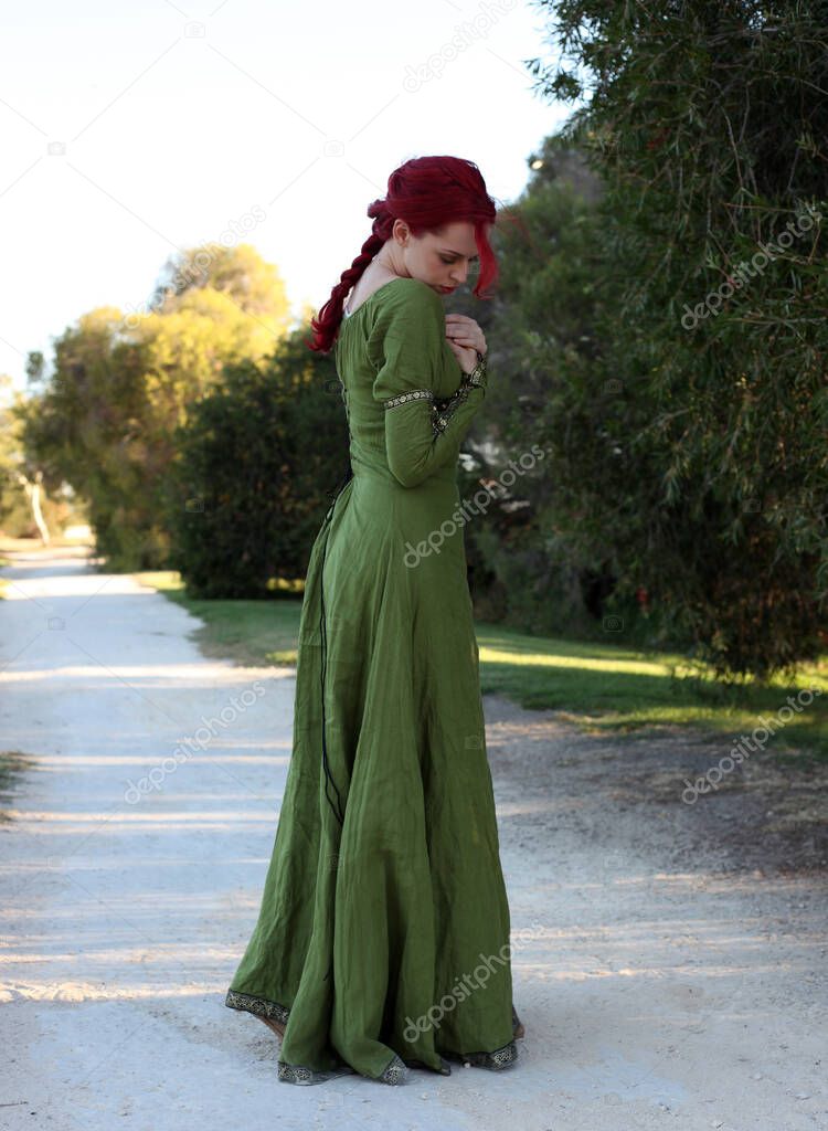 Full length portrait of red haired woman wearing a  beautiful  green medieval fantasy gown. Posing with gestural hands on a enchanted forest background.