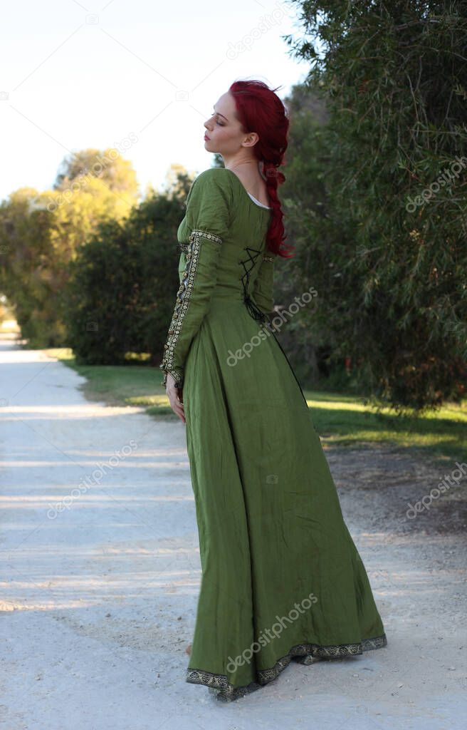 Full length portrait of red haired woman wearing a  beautiful  green medieval fantasy gown. Posing with gestural hands on a enchanted forest background.