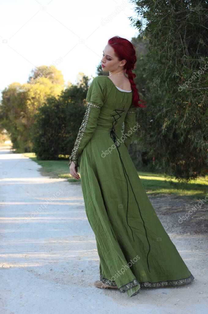 Full length portrait of red haired woman wearing a  beautiful  green medieval fantasy gown. Posing with gestural hands on a enchanted forest background.