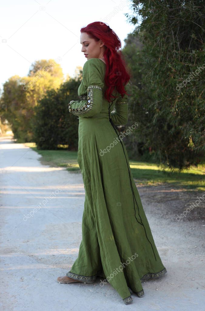 Full length portrait of red haired woman wearing a  beautiful  green medieval fantasy gown. Posing with gestural hands on a enchanted forest background.