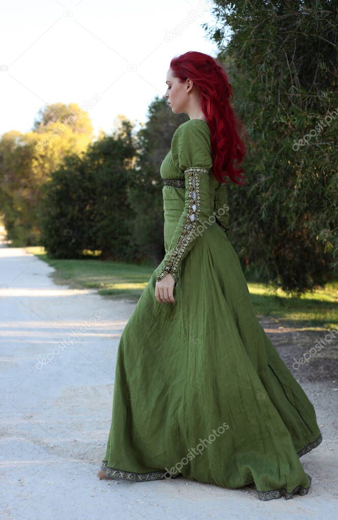 Full length portrait of red haired woman wearing a  beautiful  green medieval fantasy gown. Posing with gestural hands on a enchanted forest background.