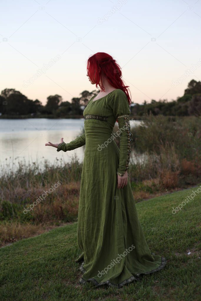 Full length portrait of red haired woman wearing a  beautiful  green medieval fantasy gown. Posing with gestural hands on a enchanted forest background.