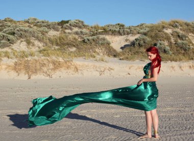 Yırtık gemi enkazı giymiş kızıl saçlı kadının tam boy portresi. Kayalık okyanus sahili arka planında el hareketleriyle poz vermek.