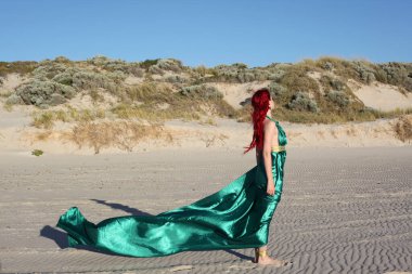 Yırtık gemi enkazı giymiş kızıl saçlı kadının tam boy portresi. Kayalık okyanus sahili arka planında el hareketleriyle poz vermek.