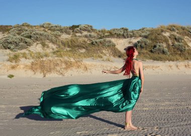 Yırtık gemi enkazı giymiş kızıl saçlı kadının tam boy portresi. Kayalık okyanus sahili arka planında el hareketleriyle poz vermek.