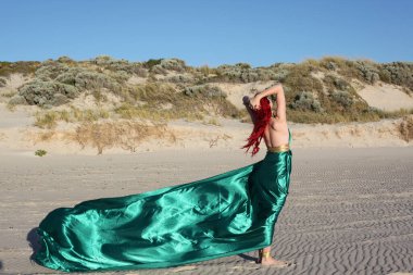 Yırtık gemi enkazı giymiş kızıl saçlı kadının tam boy portresi. Kayalık okyanus sahili arka planında el hareketleriyle poz vermek.