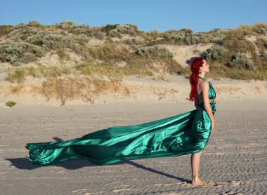 Yırtık gemi enkazı giymiş kızıl saçlı kadının tam boy portresi. Kayalık okyanus sahili arka planında el hareketleriyle poz vermek.