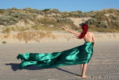 Yırtık gemi enkazı giymiş kızıl saçlı kadının tam boy portresi. Kayalık okyanus sahili arka planında el hareketleriyle poz vermek.