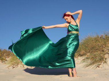 Yırtık gemi enkazı giymiş kızıl saçlı kadının tam boy portresi. Kayalık okyanus sahili arka planında el hareketleriyle poz vermek.