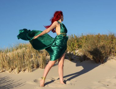 Yırtık gemi enkazı giymiş kızıl saçlı kadının tam boy portresi. Kayalık okyanus sahili arka planında el hareketleriyle poz vermek.