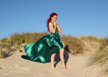 Yırtık gemi enkazı giymiş kızıl saçlı kadının tam boy portresi. Kayalık okyanus sahili arka planında el hareketleriyle poz vermek.