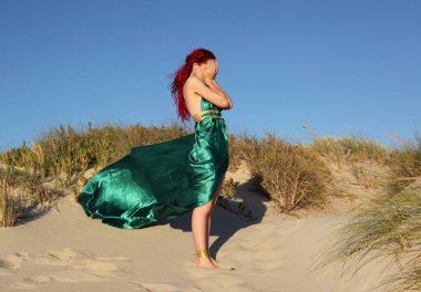 Yırtık gemi enkazı giymiş kızıl saçlı kadının tam boy portresi. Kayalık okyanus sahili arka planında el hareketleriyle poz vermek.