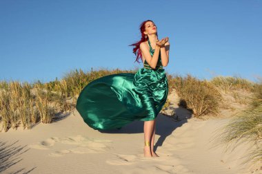 Yırtık gemi enkazı giymiş kızıl saçlı kadının tam boy portresi. Kayalık okyanus sahili arka planında el hareketleriyle poz vermek.