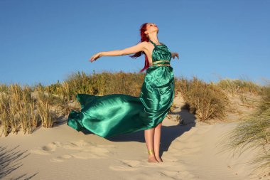 Yırtık gemi enkazı giymiş kızıl saçlı kadının tam boy portresi. Kayalık okyanus sahili arka planında el hareketleriyle poz vermek.