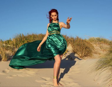 Yırtık gemi enkazı giymiş kızıl saçlı kadının tam boy portresi. Kayalık okyanus sahili arka planında el hareketleriyle poz vermek.