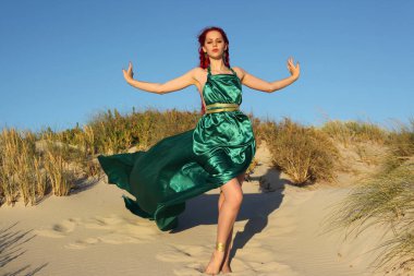 Yırtık gemi enkazı giymiş kızıl saçlı kadının tam boy portresi. Kayalık okyanus sahili arka planında el hareketleriyle poz vermek.