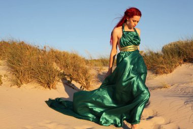 Yırtık gemi enkazı giymiş kızıl saçlı kadının tam boy portresi. Kayalık okyanus sahili arka planında el hareketleriyle poz vermek.