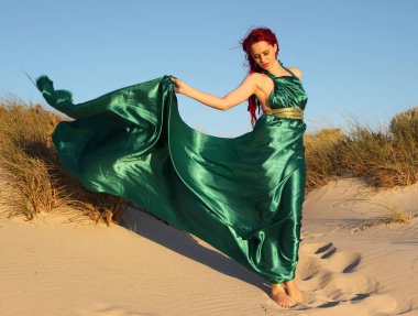 Yırtık gemi enkazı giymiş kızıl saçlı kadının tam boy portresi. Kayalık okyanus sahili arka planında el hareketleriyle poz vermek.