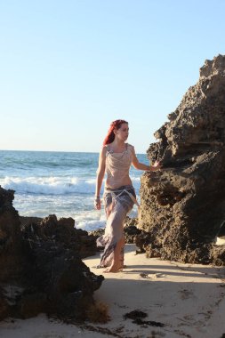 Yırtık gemi enkazı giymiş kızıl saçlı kadının tam boy portresi. Kayalık okyanus sahili arka planında el hareketleriyle poz vermek.