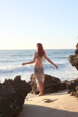 Yırtık gemi enkazı giymiş kızıl saçlı kadının tam boy portresi. Kayalık okyanus sahili arka planında el hareketleriyle poz vermek.