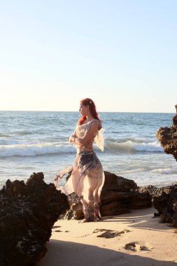 Yırtık gemi enkazı giymiş kızıl saçlı kadının tam boy portresi. Kayalık okyanus sahili arka planında el hareketleriyle poz vermek.