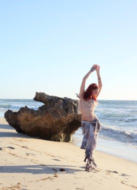 Yırtık gemi enkazı giymiş kızıl saçlı kadının tam boy portresi. Kayalık okyanus sahili arka planında el hareketleriyle poz vermek.