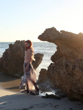 Yırtık gemi enkazı giymiş kızıl saçlı kadının tam boy portresi. Kayalık okyanus sahili arka planında el hareketleriyle poz vermek.