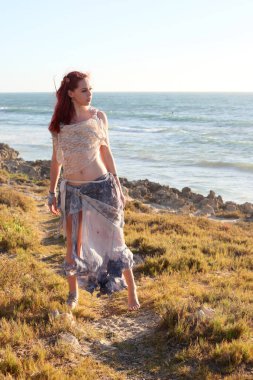 Yırtık gemi enkazı giymiş kızıl saçlı kadının tam boy portresi. Kayalık okyanus sahili arka planında el hareketleriyle poz vermek.