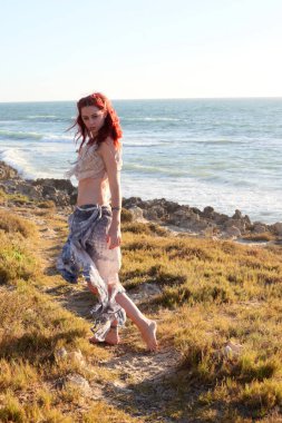 Yırtık gemi enkazı giymiş kızıl saçlı kadının tam boy portresi. Kayalık okyanus sahili arka planında el hareketleriyle poz vermek.