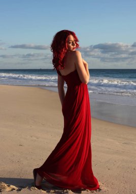 Kızıl saçlı, zarif kırmızı bir elbise giymiş bir kadının tam boy portresi. Gün batımında, okyanus sahili manzarasında el hareketleriyle poz vermek..