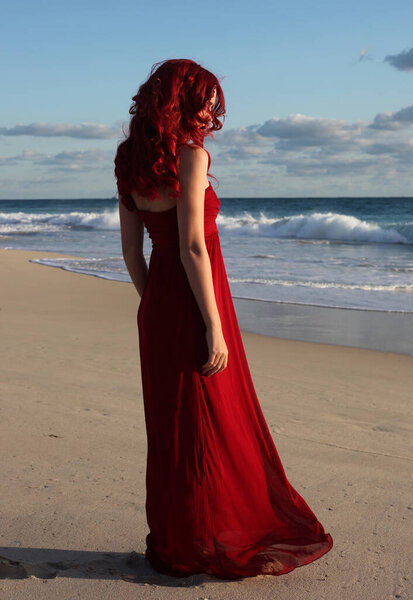 Полный портрет рыжеволосой женщины в элегантном красном платье. Стоя поза с жестом руки на закате океана пейзаж пляжа.