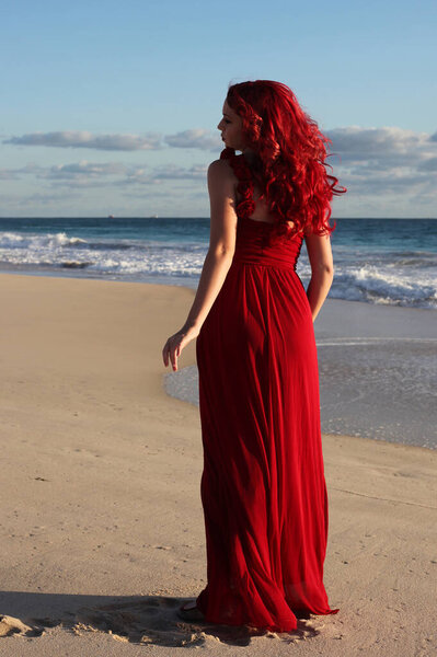 Полный портрет рыжеволосой женщины в элегантном красном платье. Стоя поза с жестом руки на закате океана пейзаж пляжа.