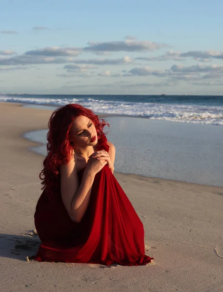 Full length portrait of  redhead woman wearing elegant red gown. Standing  pose with gestural hands at sunset ocean beach landscape background.