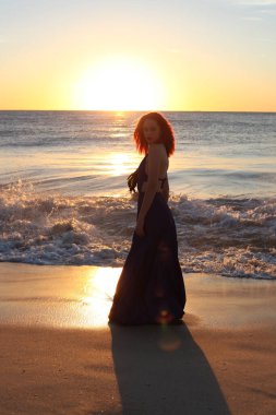 Zarif bir elbise giyen kızıl saçlı kadının tam boy portresi. Gün batımında, okyanus sahili manzarasında el hareketleriyle poz vermek..