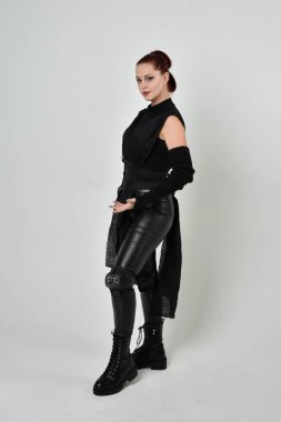 Siyah fütürist bilim kurgu deri kostüm giyen güzel kızıl saçlı kadın modelin tam boy portresi. Dinamik duruş, el hareketleriyle beyaz bir stüdyonun arka planında..